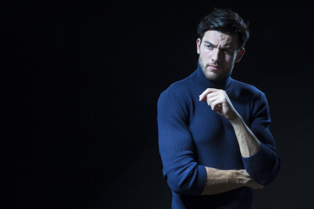 白人英俊的黑发男子的肖像与微笑摆姿势与复制空间的黑色背景。穿着蓝色高领毛衣，双手合十，摆出一个成功青年的造型图片下载