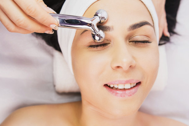 上面的照片，美丽的快乐的女人得到脸按摩辊设备由美容师在美容诊所沙龙。白人黑发女性病人接受举重治疗图片下载