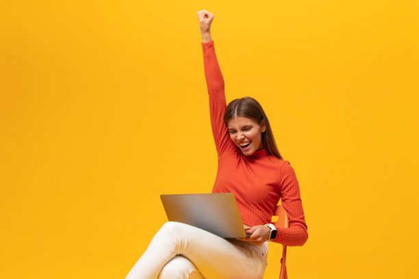 赢家。年轻女子兴奋地坐在椅子上，拿着笔记本电脑，喊着“是”，孤立在黄色上图片下载