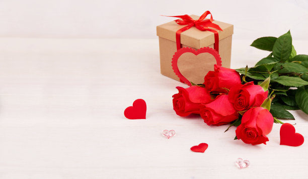 一束红玫瑰，一个牛皮纸包装的盒子，用红色的丝带和心形装饰在白色的木质背景上。情人节贺卡，母亲节贺卡，生日贺卡。图片下载