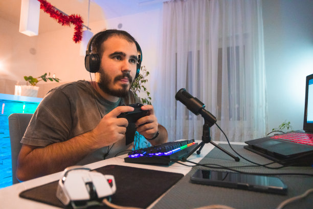 专注的年轻男性玩家身体前倾，戴着耳机，手持操纵杆控制器，对着麦克风讲话，从家里直播图片下载