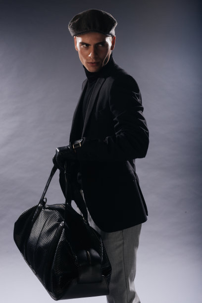 这张照片是一个下巴很尖的年轻人，头戴报童帽，手里拿着一个男包，在工作室里以白色为背景摆姿势。图片下载