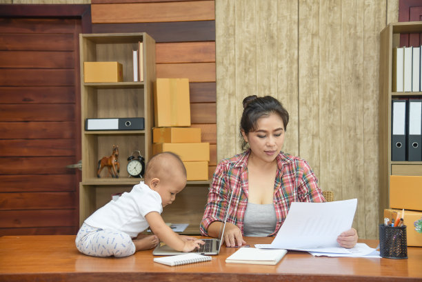 年轻漂亮的单身妈妈在家工作，拿着文件，小女儿在桌子上玩笔记本电脑。图片下载