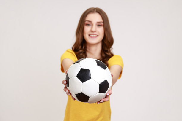 漂亮的棕色头发的年轻女性，微笑着看着镜头，提供足球，要求踢足球比赛。图片下载