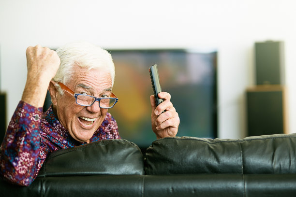一位快乐的老人坐在电视机前，手里拿着遥控器，高兴地挥舞着拳头图片下载
