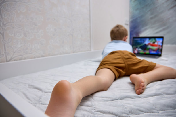 一个戴着无线耳机的金发小男孩在床上用着一台现代化的笔记本电脑。图片下载