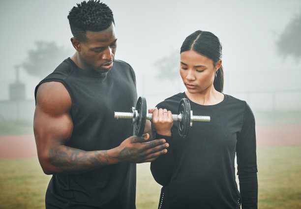 这张照片拍摄的是一个运动的年轻女子，在训练师的帮助下，在户外用哑铃锻炼身体图片下载