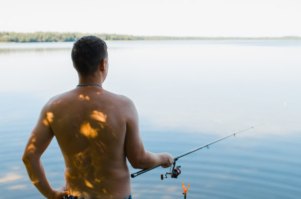 体育及爱好、休闲娱乐观念。男渔夫夏日清晨户外垂钓，后视镜。一名白人男子手持旋转棒站在自然的湖滨，齐腰图片下载