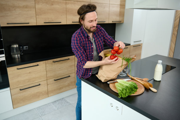 一个快乐的年轻人站在家里的厨房里，从一个环保纸袋里拿出蔬菜和健康食品，手里拿着一根新鲜的番茄，放在厨房的桌子上图片下载