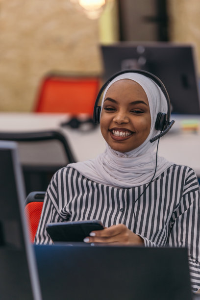 一个戴着头巾和耳机的非洲女孩在一个现代化的办公室里做客户支持。图片下载