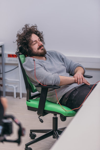 一头卷发的男员工睡在办公室的椅子上。图片下载