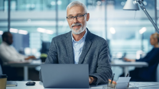 现代办公室:成功中年大胡子商人的肖像，在办公桌前用笔记本电脑工作，面带微笑。多元化的工作场所与专业人士。图片下载