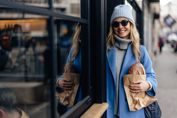 一个微笑的女人，穿着蓝色的外套和帽子，靠在咖啡馆的门上。图片下载