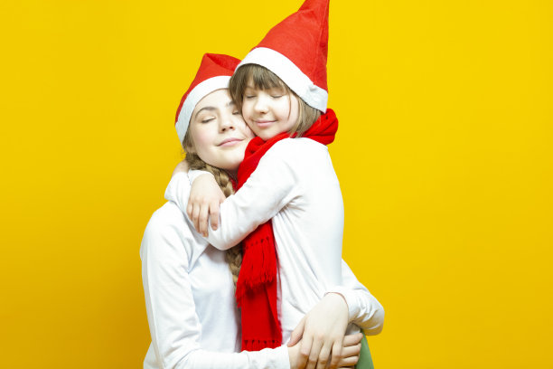两个白种人金发姐妹在冬季圣诞老人的帽子拥抱拥抱在一起在黄色的背景白色休闲服装图片下载