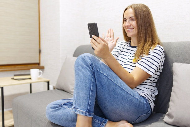 一名兴高采烈的年轻女子正在用智能手机打视频电话，用手势向朋友或父母打招呼。年轻的白人妇女微笑着放松地坐在客厅的沙发上进行视频通话图片下载