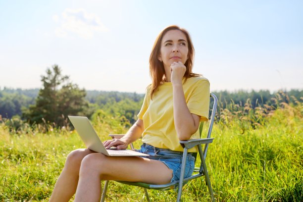 暑假在野外草地上，成年女子坐在折叠椅上用笔记本电脑露营图片下载