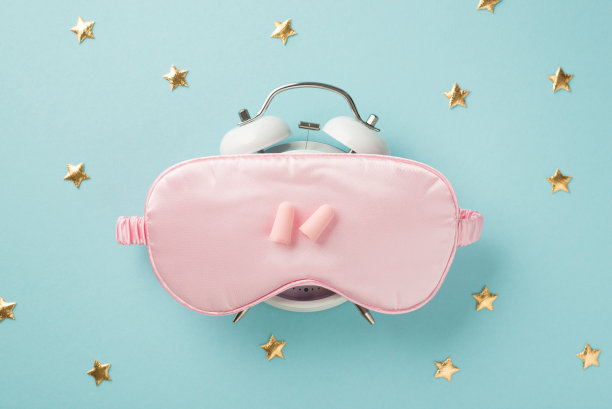 顶视图照片的金色星星耳塞和粉红色丝绸睡眠面具闹钟上孤立的淡蓝色背景与空空间图片下载