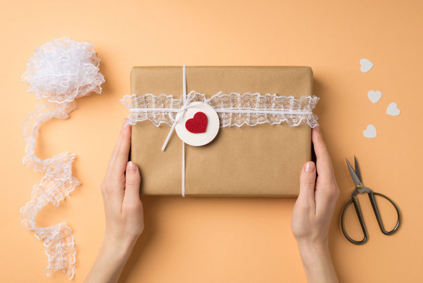 第一人称顶视图照片的情人节装饰女人的手拿牛皮纸礼品盒与花边装饰标签与红色心形剪刀标签线轴的蕾丝在孤立的米色背景图片下载