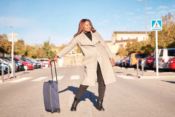 快乐的非洲裔美国人，穿着黑色裤子和米色夹克，拿着一个手提箱，在机场附近摆姿势。图片下载