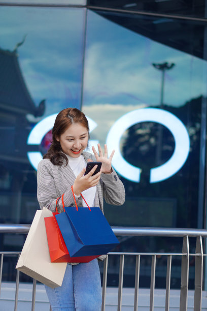 一位满面笑容的亚洲少女站在购物中心前，手里拿着彩色购物袋和手机。生活方式的技术概念图片下载