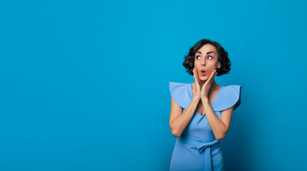 这张照片拍摄的是一个美丽的女人，她穿着一件蓝色的时髦连衣裙，她抱着自己的脸颊，目光移开，孤立在蓝色的背景上图片下载