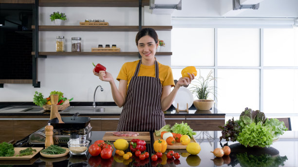 年轻的家庭主妇站在那里微笑着，双手拿着红椒和黄椒。厨房柜台上摆满了各种各样的蔬菜。图片下载