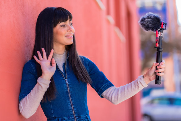 黑发女孩用手机和麦克风录制视频博客，身穿蓝色牛仔套装用于社交网络。白种人漂亮的红色背景图片下载