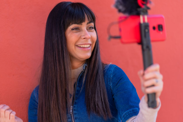 黑发女孩微笑着用手机和麦克风录制视频博客，身穿蓝色牛仔套装，为社交网络服务。白种人漂亮的红色背景图片下载