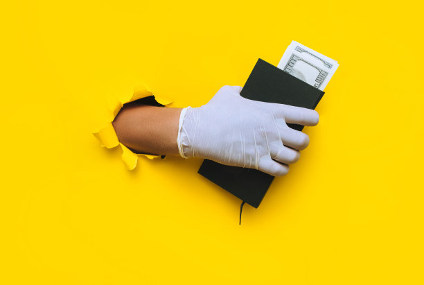 戴着白色乳胶医用手套的医生的一只手拿着一本黄色背景上印着美钞的黑色笔记本。贿赂的概念，给黑人外科医生的报酬图片下载