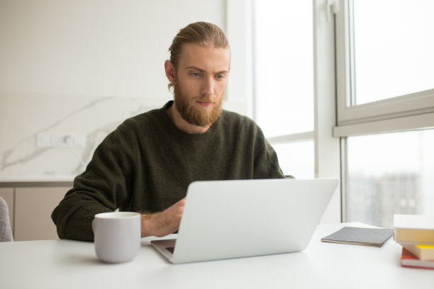 年轻人拿着杯子坐在桌子旁，在家里用笔记本电脑工作图片下载