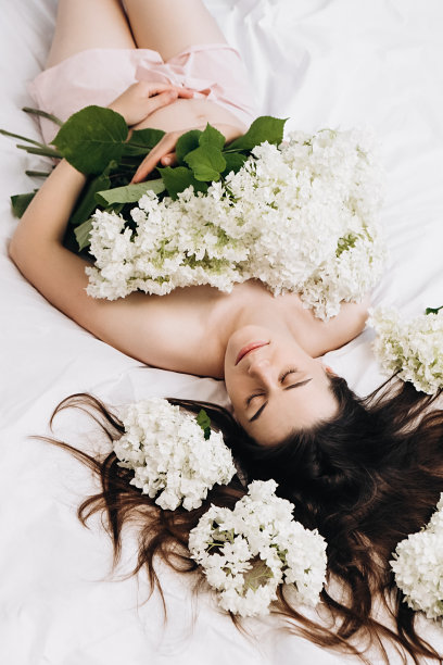 年轻孕妇的头像照片，完美的皮肤轻轻抚摸腹部的手躺在舒适的白色床上，美丽的春天鲜花。母亲的概念。未来母亲的期望图片下载