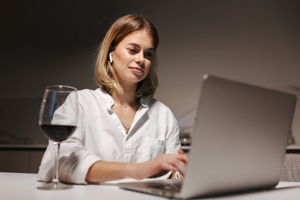 戴着耳机的年轻女士坐在家里附近，喝着红酒，在笔记本电脑上工作图片下载