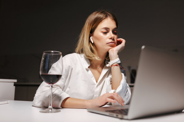 戴着耳机，沉思的女士坐在家里附近的黑暗中，拿着一杯红酒，在笔记本电脑上工作图片下载