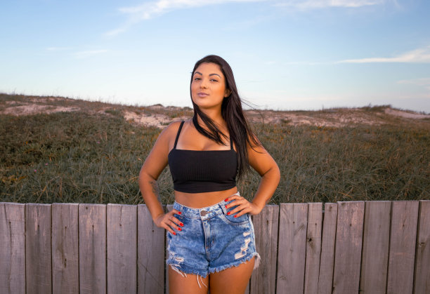 黑发模特站在海滩木板路严肃的表情和态度。身穿蓝色牛仔短裤和黑色七分裤的蓝色调照片。提到假期和放松。图片下载