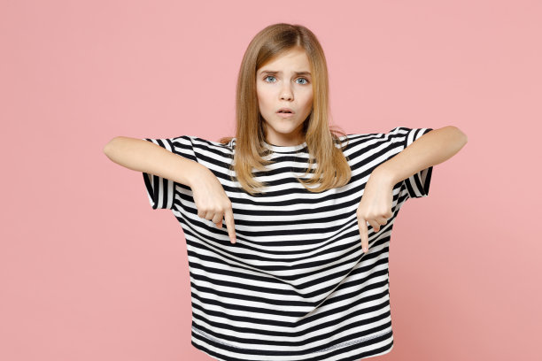 小金发微笑的小女孩12-13岁，穿着条纹的超大t恤，食指落在拷贝空间区域孤立在淡粉色背景的孩子。童年的生活方式的概念。图片下载
