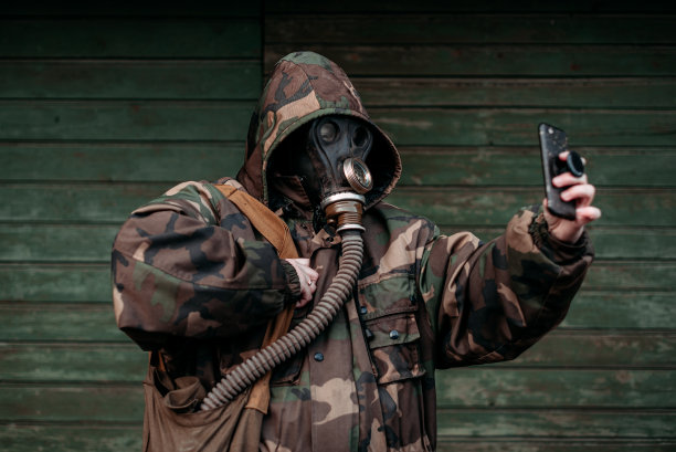 一名戴着旧防毒面具、身穿军装、头戴兜帽的妇女用手机自拍。概念-核裁军和辐射防护，流行病，冠状病毒，covid-19，大流行图片下载