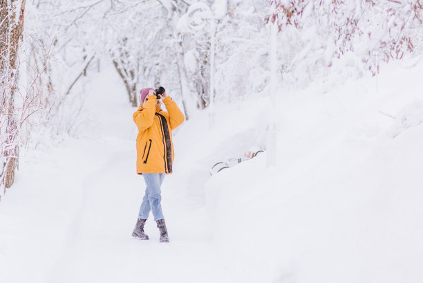 美丽的女孩穿着黄色夹克摄影师在冬季公园拍摄雪的照片图片下载