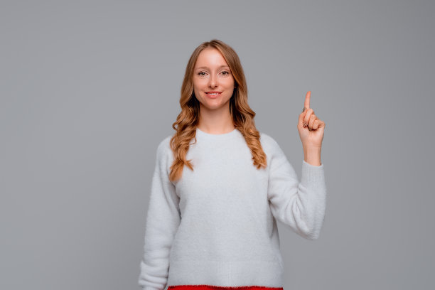 一个金发女人开心地笑着，竖起手指，责骂或斥责某人，说:请注意，穿白色毛衣，灰色背景图片下载