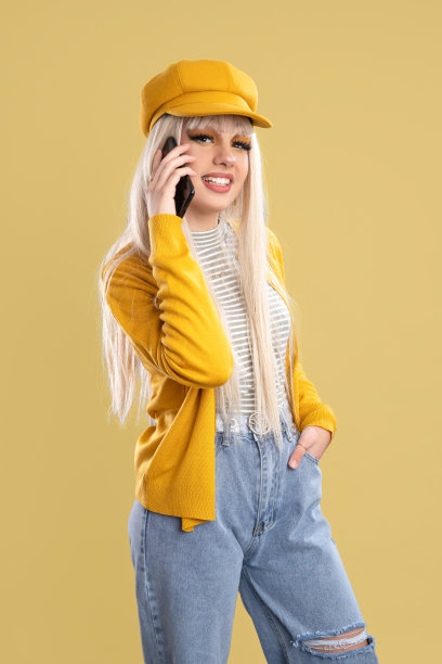 戴着帽子，穿着黄色夹克的金发女人在黄色背景下讲电话图片下载