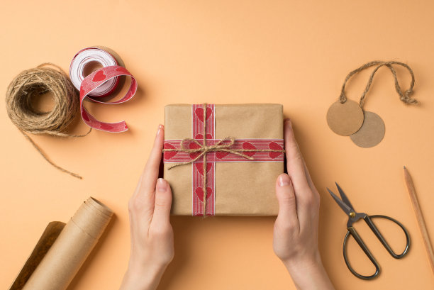 第一人称俯视图照片的情人节装饰女性的手握工艺纸礼品盒手工工具剪刀卷牛皮纸卷筒丝带缠绕铅笔在孤立的米色背景图片下载