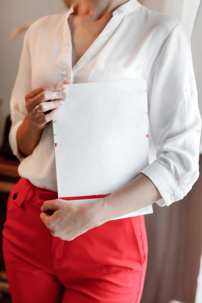 裁剪照片的年轻女子拿着一本杂志与模拟。穿白衬衫红裤子的女孩站在窗边图片下载