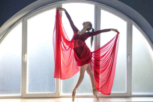 美丽的年轻芭蕾舞女演员穿着优雅的红色芭蕾长裙，摆出蝴蝶的姿势，站在芭蕾舞工作室的大圆形窗户前，自然光线通过环境光照亮室内图片下载