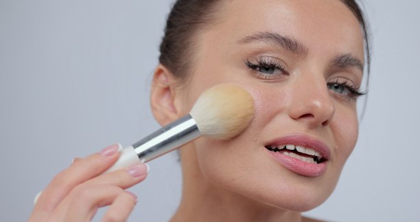 年轻的女人用刷子在她的脸上化妆腮红，而站在工作室的灰色背景。概念美容、护肤。图片下载