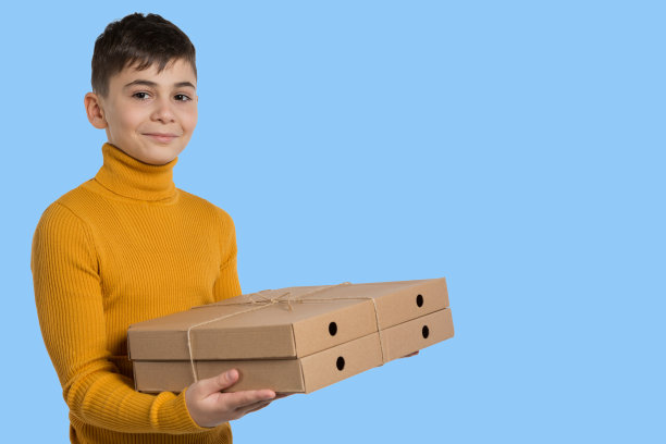 一个穿着黄色毛衣的男孩微笑着，拿着两个披萨盒，上面是送外卖的，背景是浅蓝色图片下载