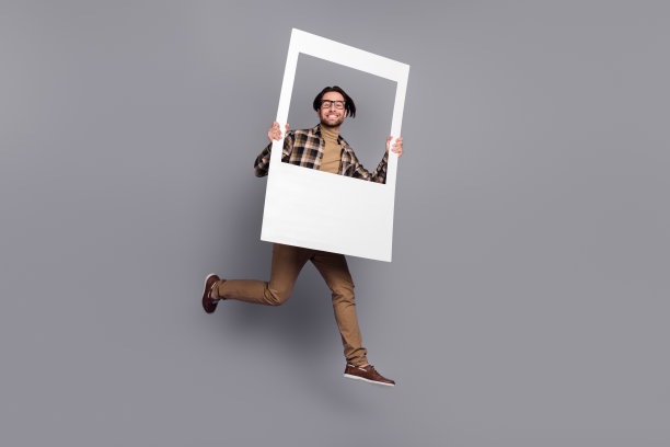 滑稽的男子照片，穿格子衬衫，眼镜跳高，持有白色框架孤立的灰色背景图片下载