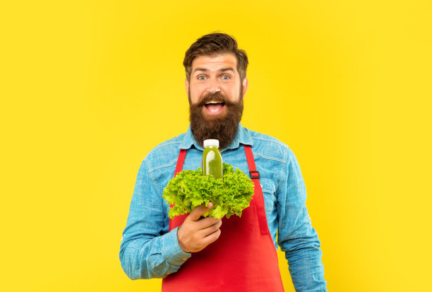 穿着围裙的男子惊讶地拿着黄色背景的新鲜生菜和果汁瓶，果汁酒保图片下载