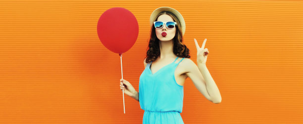美丽的年轻女子的肖像吹着她的嘴唇送空中的吻红色气球，戴着一顶夏季草帽在橙色的背景图片下载