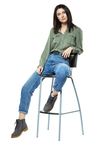 一个穿着牛仔裤、靴子和卡其色衬衫的年轻女子正坐在一把高椅上。孤立在白色的。图片下载