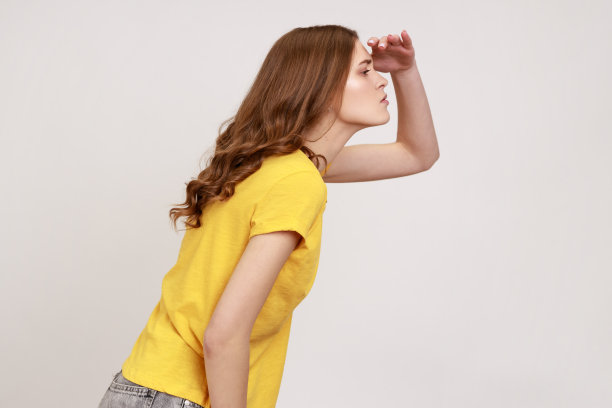 美丽的波浪式头发的年轻女性在黄色t恤看远与手以上的眼睛，期待未来。图片下载