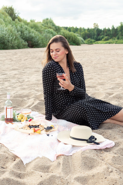 迷人的年轻女子拿着一杯玫瑰葡萄酒在沙滩上。玫瑰葡萄酒，各种水果，面包和奶酪图片下载